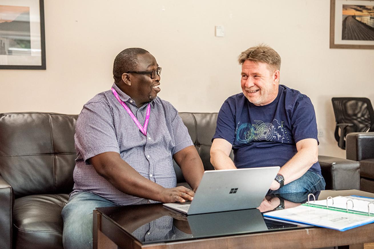 A supported service user and a member of staff sat together and smiling, they are sat on a brown leather sofa, in front of a laptop on top of a coffee table.