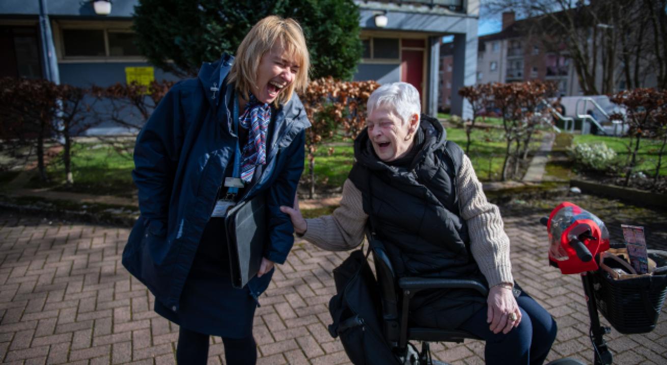 A woman sitting in a mobility scooter leaning back and laughing with a woman that is standing up, holding a clipboard and wearing a Sanctuary lanyard