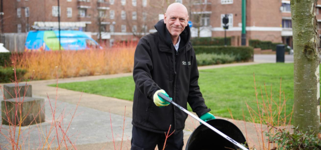 A man wearing black trousers and a black Sanctuary jacket wearing gloves a picking up litter on an estate