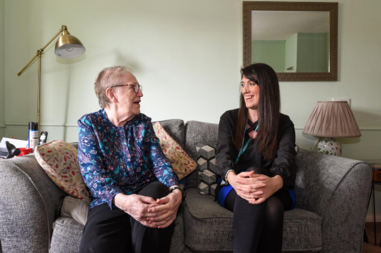 An elderly woman wearing a blue flowery top and black trousers sitting on a grey sofa smiling with a younger woman wearing a black suit and a Sanctuary lanyard