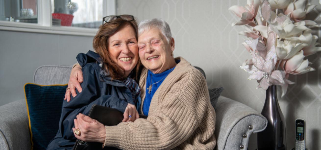 Two women smiling and hugging each other whilst sitting on a grey sofa