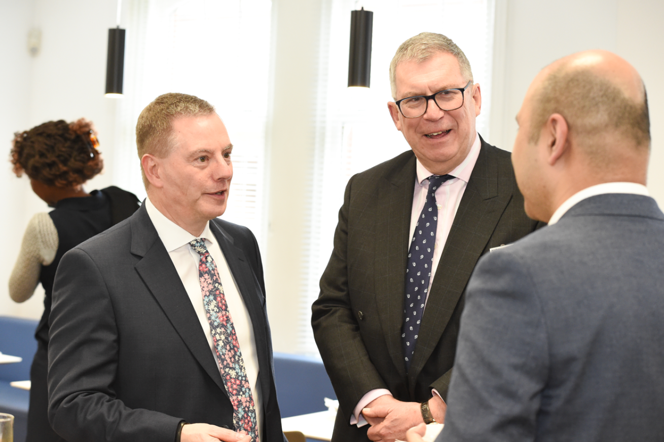 Three men wearing suits standing talking to each other in a bright room with blue seating