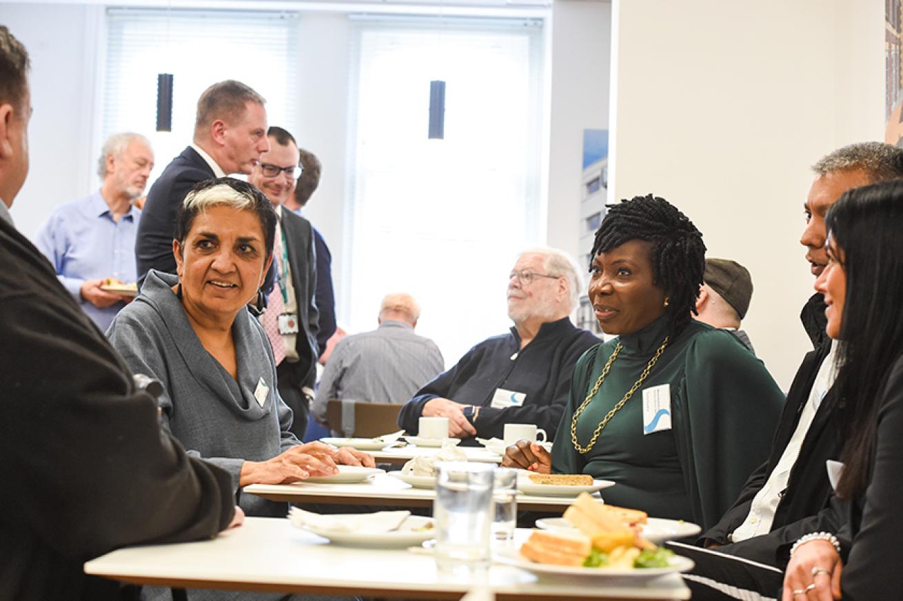 Members of Sanctuary's boards standing and sitting in a canteen with plates of food on the tables.