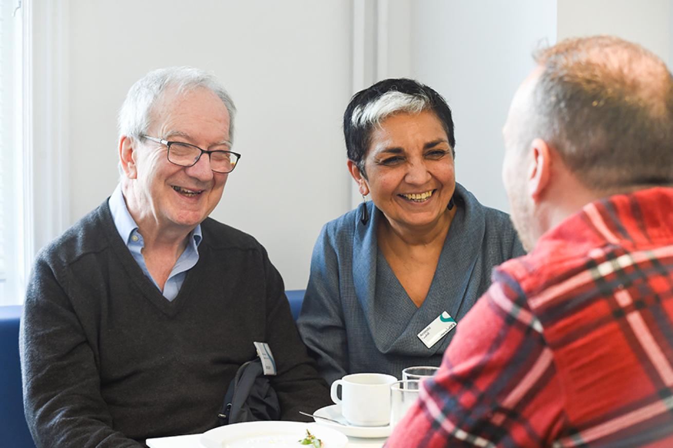 A woman with black and white hair wearing a grey top sitting on a blue bench next to a man wearing a  blue shirt and grey jumper. Both people are wearing Sanctuary name badges and smiling at a man wearing a red checked shirt.