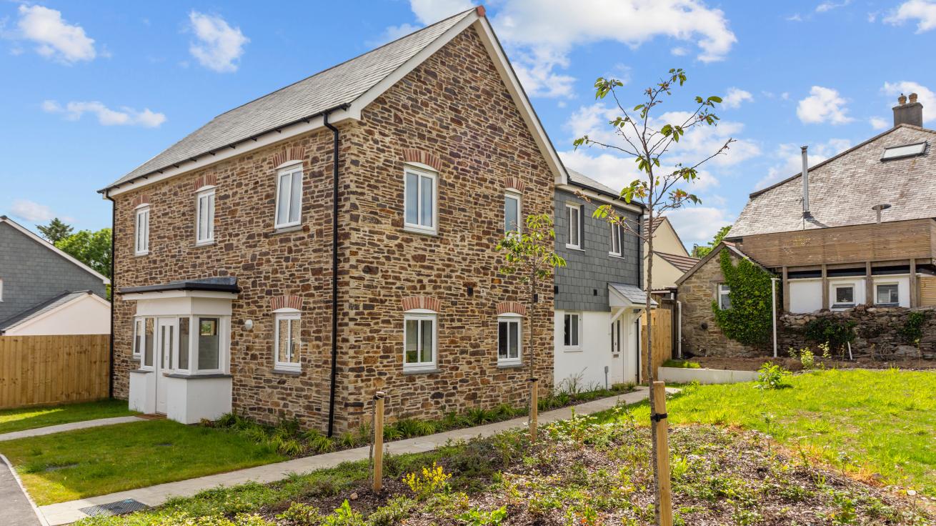 Exterior of a detached stone brick house with a grey roof and a newly planted tree in the foreground