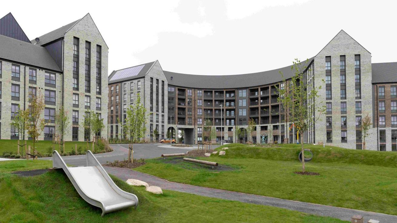 Exterior of blocks of grey brick buildings with black and brown accents. There is a grassy area in the foreground with trees and children's playground equipment located on it.