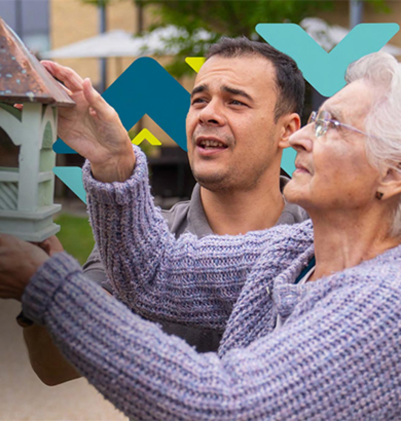 An elderly woman is helped by a younger man to hang a green bird house on a wooden wall