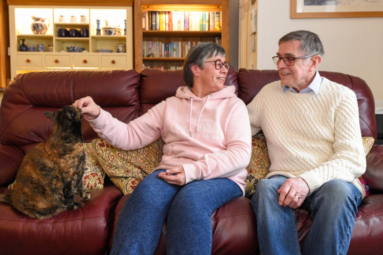 An elderly woman and man sitting on a burgundy leather sofa in a family living room. The woman is feeding an orange and black cat