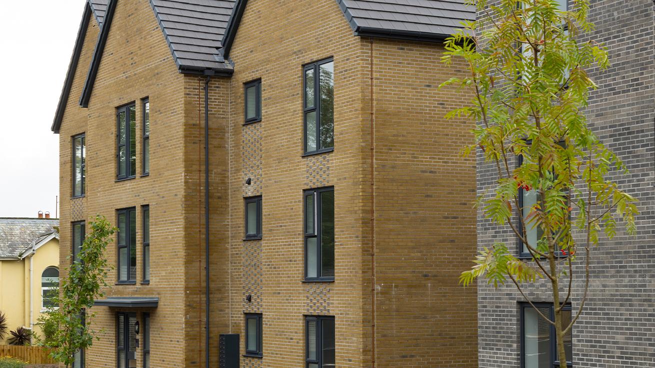 Exterior of a brown brick building next to a grey brick building. Both buildings have black accents with trees planted in front