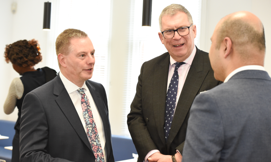 Three men wearing suits standing talking to each other in a bright room with blue seating