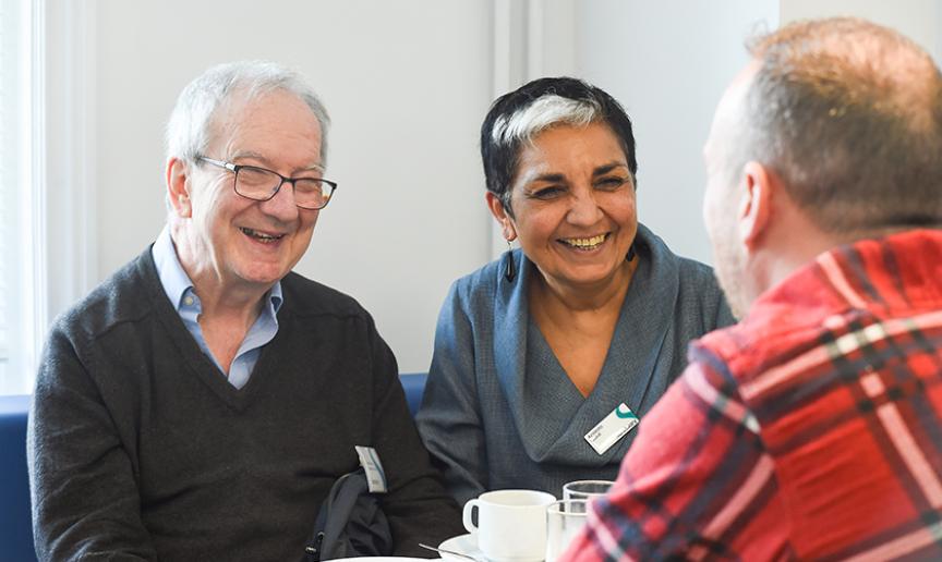 A woman with black and white hair wearing a grey top sitting on a blue bench next to a man wearing a  blue shirt and grey jumper. Both people are wearing Sanctuary name badges and smiling at a man wearing a red checked shirt.