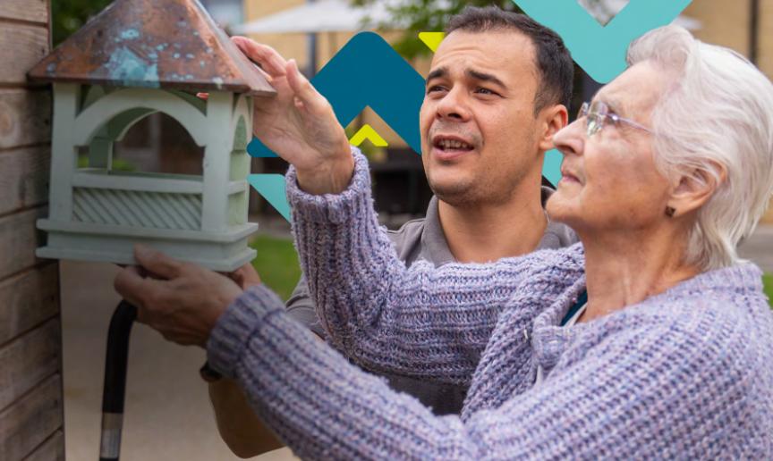 An elderly woman is helped by a younger man to hang a green bird house on a wooden wall