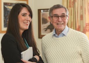 A white woman with black hair wearing a black jacket and holding a notepad stood next to an white older man wearing a cream jumper