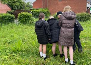 A woman and school children standing in a greasy field with redbrick houses in the background